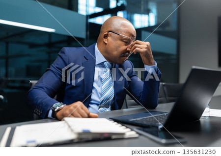 Tired black mature businessman rubbing eyes at desk while working on laptop 135631230