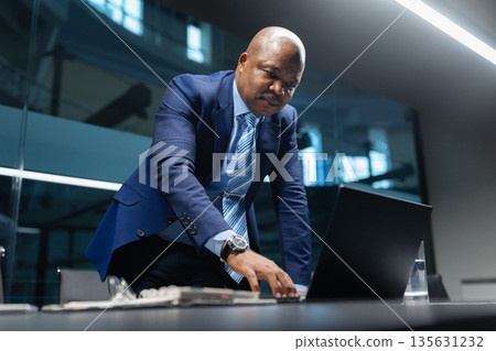 Focused black mature businessman leaning over conference table while working on laptop Focused black mature businessman leaning over conference table while working on laptop 135631232