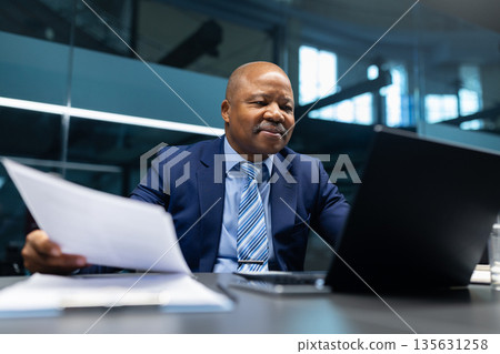 Confident black mature businessman reviewing paperwork while sitting at desk with laptop Confident black mature businessman reviewing paperwork while sitting at desk with laptop 135631258