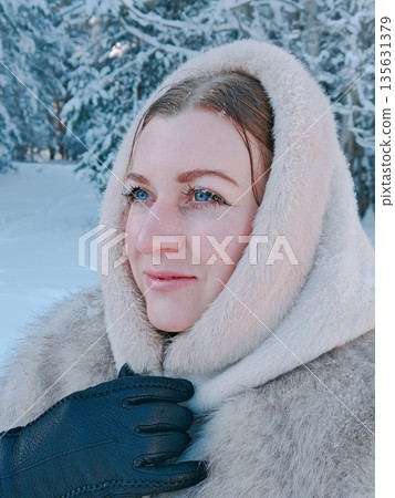 Young Woman Portrait in Snowy Winter Forest Close-Up. Close-up portrait of joyful woman with snow on eyelashes in winter forest, wearing fluffy fur coat. Vertical lifestyle, serene winter moment. 135631379