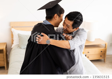 Graduation celebration, father hugging son in cap and gown at home 135631515
