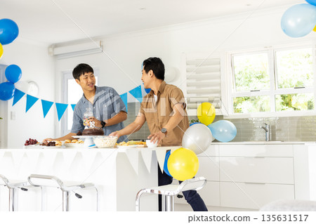 Two men preparing birthday celebration in kitchen with balloons and cake Two men preparing birthday celebration in kitchen with balloons and cake 135631517