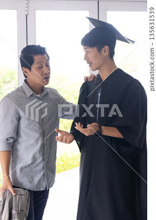 Proud father congratulating son in graduation gown, sharing joyful moment at home 135631539