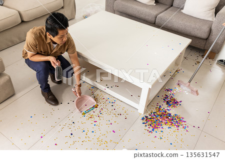 Man cleaning colorful confetti from floor with dustpan and broom at home 135631547