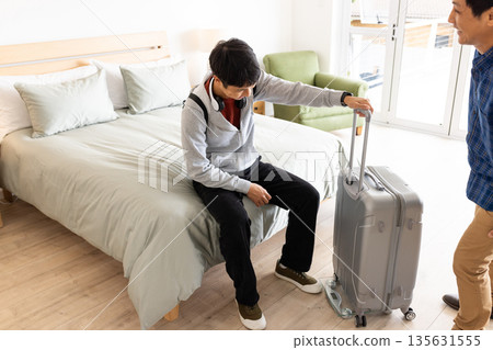 Young man sitting on bed with suitcase, preparing for travel at home 135631555