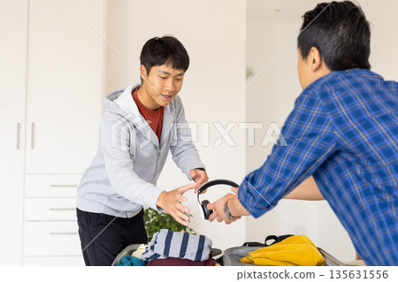 Young man packing suitcase with headphones, preparing for travel with friend at home 135631556