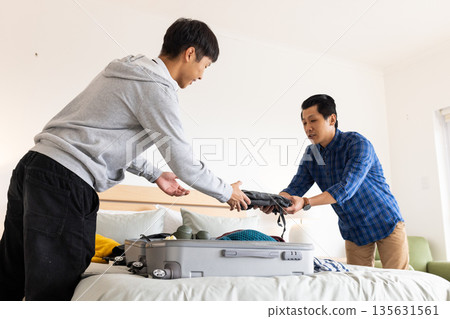 Father and son packing suitcase together on bed, preparing for travel adventure Father and son packing suitcase together on bed, preparing for travel adventure 135631561