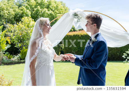 Bride and groom exchanging vows outdoors under elegant floral arch, smiling warmly 135631594