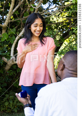 Man proposing to surprised woman in garden, holding engagement ring box 135631598