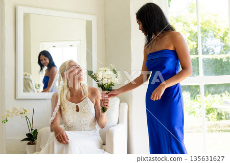 Bride holding bouquet, smiling with bridesmaid in blue dress by large window Bride holding bouquet, smiling with bridesmaid in blue dress by large window 135631627