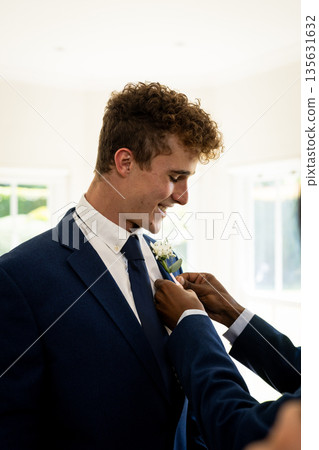 Groom smiling while friend adjusts boutonniere in bright wedding room Groom smiling while friend adjusts boutonniere in bright wedding room 135631632
