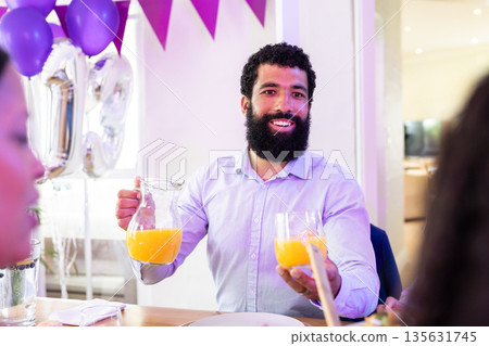 Man serving orange juice at festive gathering, smiling and engaging with guests 135631745