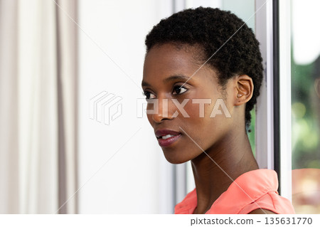 Confident woman in coral blouse looking thoughtfully out window at home 135631770