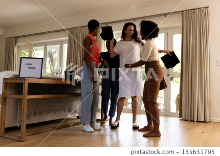 African American women celebrating school reunion at home, holding graduation caps 135631795