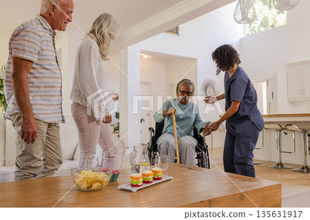 Nurse assisting senior woman in wheelchair while friends watch in living room 135631917