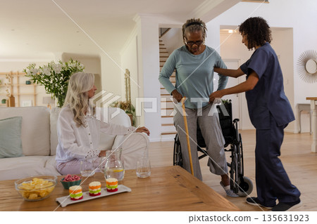 Senior woman using walking aid with caregiver while friend watches in living room Senior woman using walking aid with caregiver while friend watches in living room 135631923