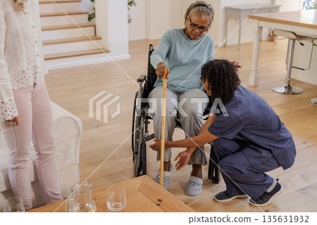 Assisting senior woman in wheelchair, African American nurse helps with walking stick 135631932