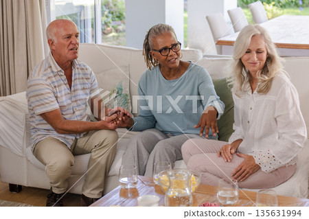 Senior friends sitting on sofa at home, enjoying conversation and holding hands 135631994