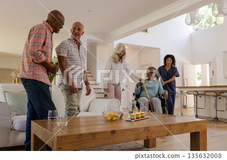 Senior friends enjoying conversation at home with caregiver assisting in wheelchair 135632008