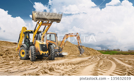 Deep tracks from heavy machinery are visible on sandy ground with a large sand pile in the background, showing active construction on a cloudy day Deep tracks from heavy machinery are visible on sandy ground with a large sand pile in the background, showing active construction on a cloudy day 135632039
