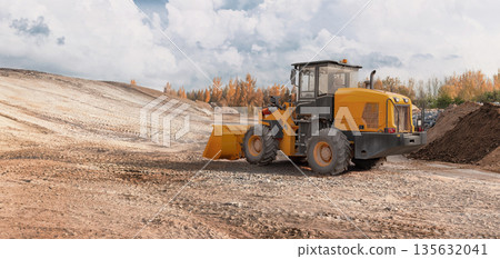 A loader is working on a construction site to level the ground. The sky is clear, and trees surround the area. Equipment is positioned on the dirt terrain 135632041