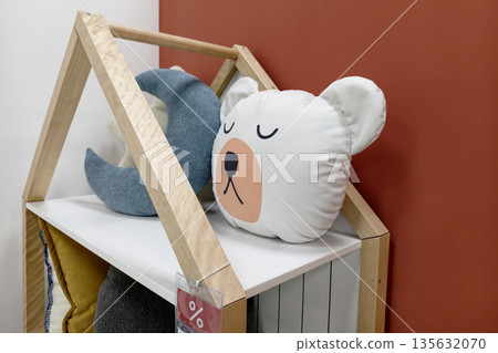 A shelf displays cushions including a bear design and a moon shape in a child's room beside a red wall during daylight hours A shelf displays cushions including a bear design and a moon shape in a child's room beside a red wall during daylight hours 135632070