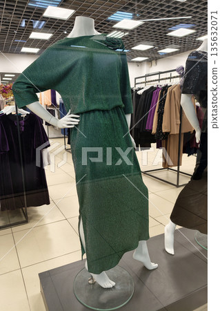 A mannequin wears a green dress positioned in a store display. Clothing racks filled with various outfits are visible in the background A mannequin wears a green dress positioned in a store display. Clothing racks filled with various outfits are visible in the background 135632071