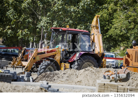 Construction equipment operates on a dirt area surrounded by trees and parked cars during the day. Workers manage tasks on the site 135632094