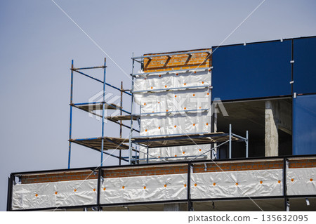 Workers continue construction on a building using scaffolding and plywood while the sun shines brightly and the sky appears clear and blue Workers continue construction on a building using scaffolding and plywood while the sun shines brightly and the sky appears clear and blue 135632095