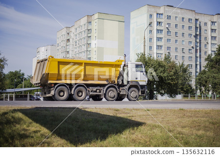 A yellow dump truck is seen moving along the road beside several tall residential buildings under a bright sky in an urban setting 135632116