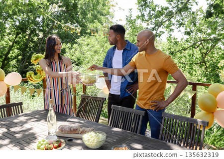Friends enjoying outdoor party, sharing snacks and smiling in sunny garden Friends enjoying outdoor party, sharing snacks and smiling in sunny garden 135632319