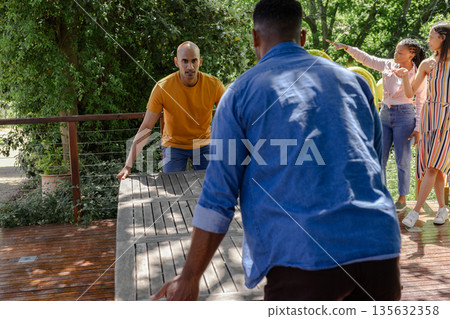 Friends setting up outdoor table for celebration, working together in sunny garden 135632358