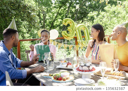 Friends celebrating birthday outdoors, woman opening gift with joy and laughter 135632387