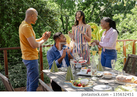 Friends celebrating birthday outdoors, clapping and laughing around decorated table Friends celebrating birthday outdoors, clapping and laughing around decorated table 135632451