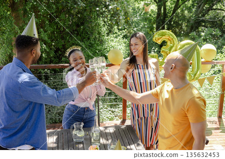 Celebrating birthday outdoors, friends toasting with champagne glasses wearing party hats 135632453