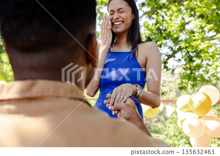 Man proposing to smiling woman outdoors, surrounded by balloons and trees Man proposing to smiling woman outdoors, surrounded by balloons and trees 135632463