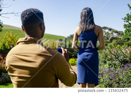 Man taking photo of woman in blue dress outdoors in garden setting 135632475