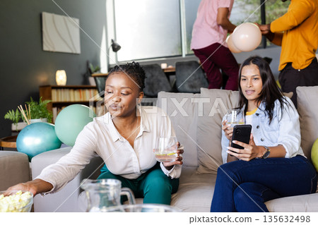 African American woman enjoying popcorn while friend uses smartphone at home party African American woman enjoying popcorn while friend uses smartphone at home party 135632498