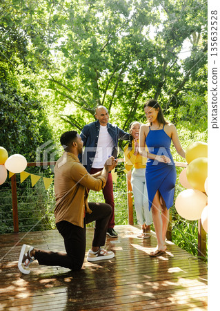 Man proposing to woman on sunny deck, friends celebrating with balloons Man proposing to woman on sunny deck, friends celebrating with balloons 135632528
