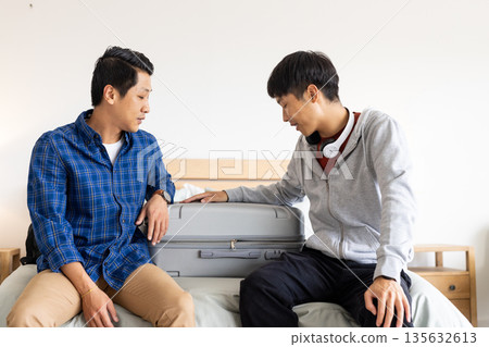 Two men sitting on bed packing suitcase, preparing for travel together Two men sitting on bed packing suitcase, preparing for travel together 135632613