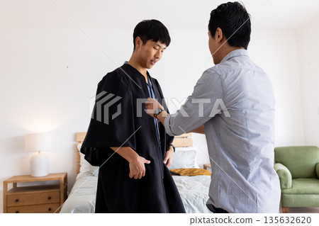 Father helping son adjust graduation gown at home, preparing for ceremony Father helping son adjust graduation gown at home, preparing for ceremony 135632620