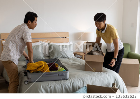 Packing suitcase and boxes, two men organizing belongings in bedroom Packing suitcase and boxes, two men organizing belongings in bedroom 135632622