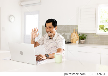Asian man video chatting on laptop in kitchen, smiling and waving hello 135632631