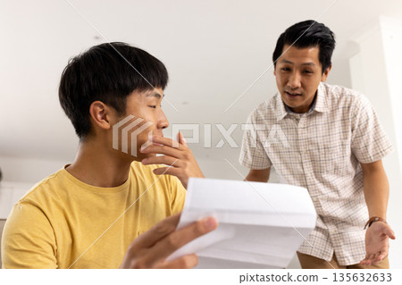 Father discussing important document with son at home, both appearing thoughtful Father discussing important document with son at home, both appearing thoughtful 135632633