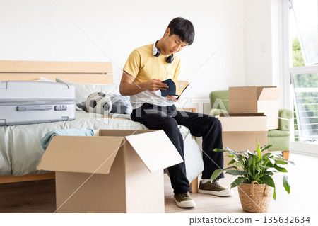 Teen unpacking boxes in bedroom, reading book with headphones around neck Teen unpacking boxes in bedroom, reading book with headphones around neck 135632634