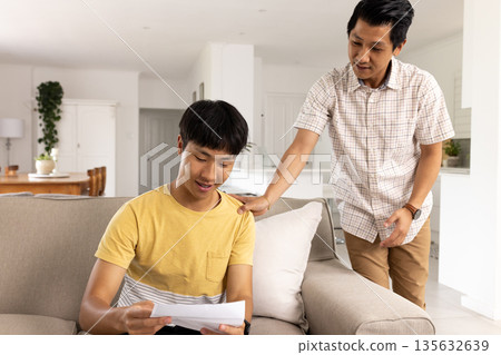 Father encouraging teenage son reading letter on couch at home, sharing proud moment Father encouraging teenage son reading letter on couch at home, sharing proud moment 135632639