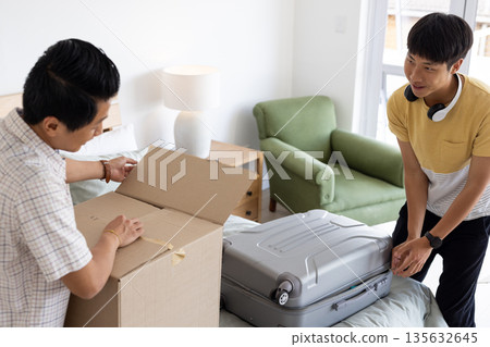 Father and son unpacking cardboard box and suitcase in bedroom, smiling together 135632645