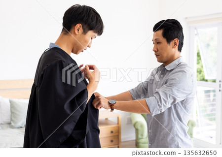 Father helping son adjust graduation gown at home, preparing for ceremony 135632650