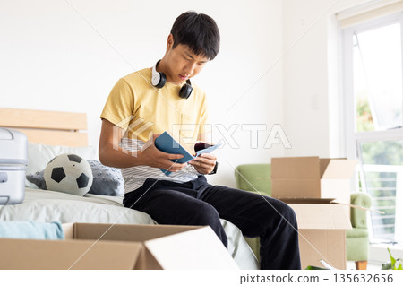 Young man reading book on bed surrounded by moving boxes and soccer ball 135632656