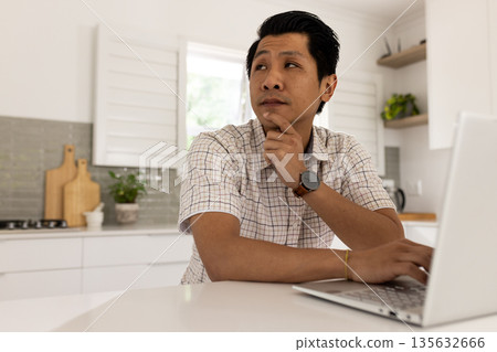 Asian man in kitchen using laptop, thinking deeply and planning work Asian man in kitchen using laptop, thinking deeply and planning work 135632666